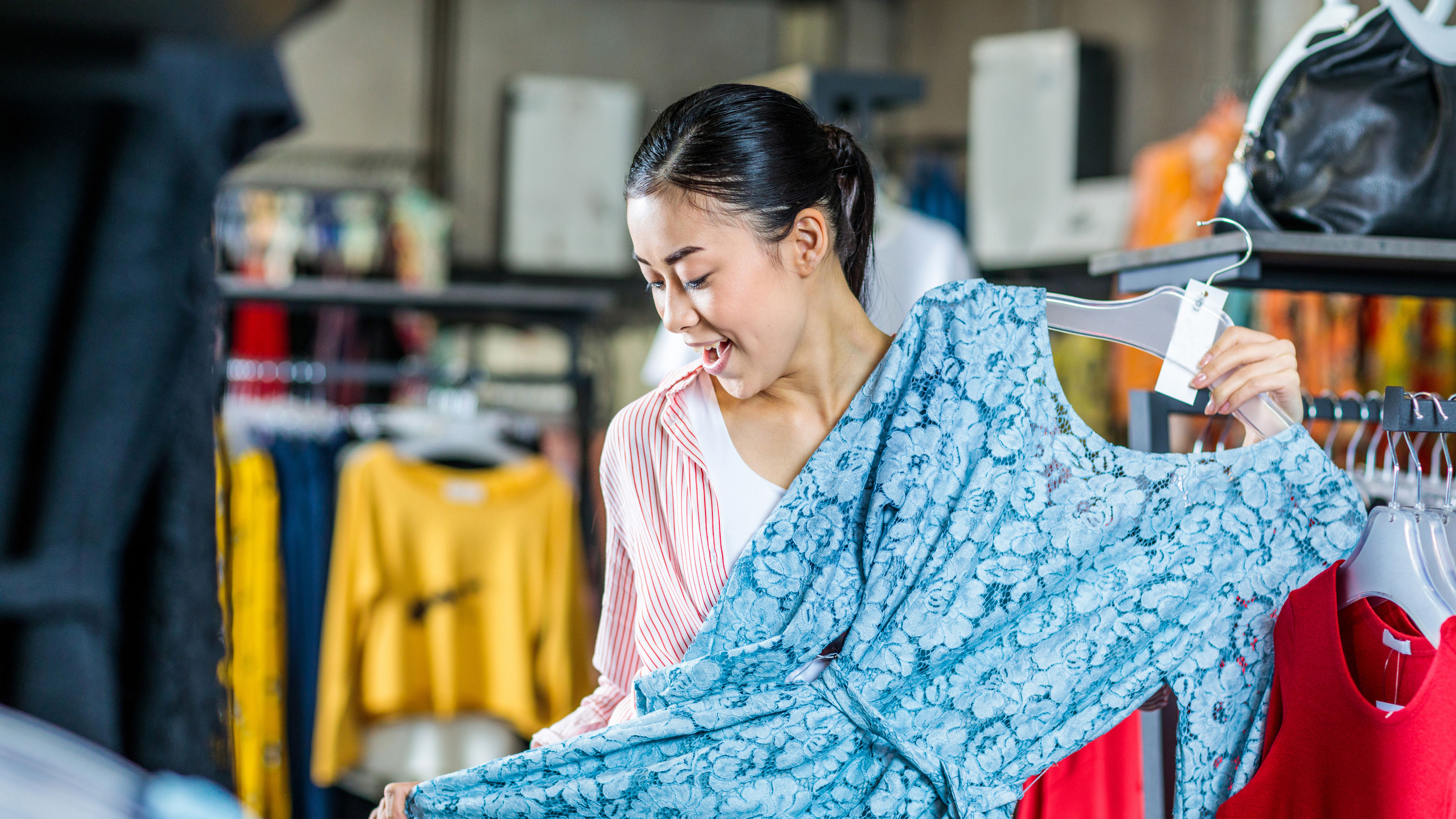 Asian girl holding a blue dress in excitement at a shop.