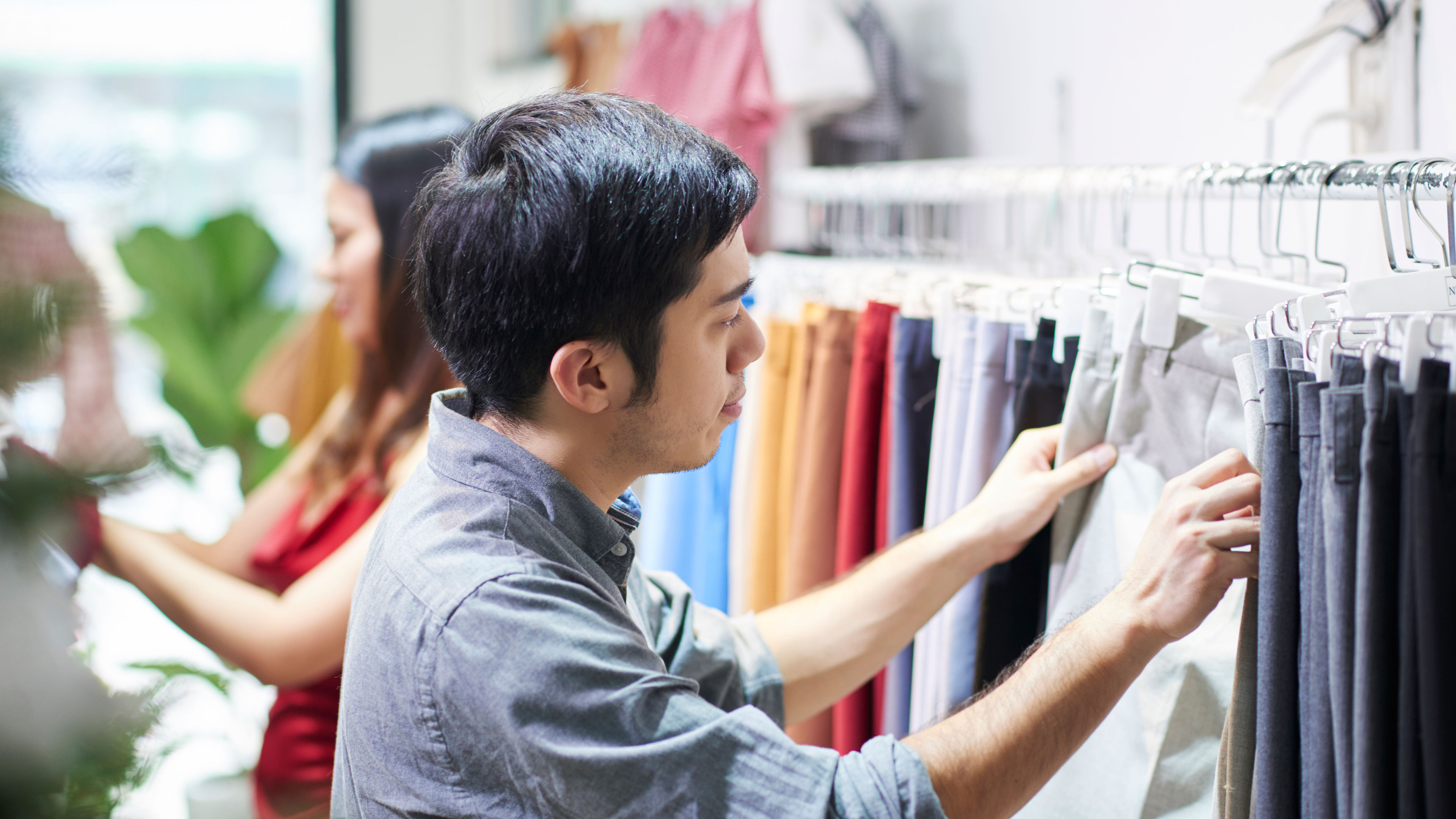 A man browsing trousers in a clothing store while a woman shops in the background.