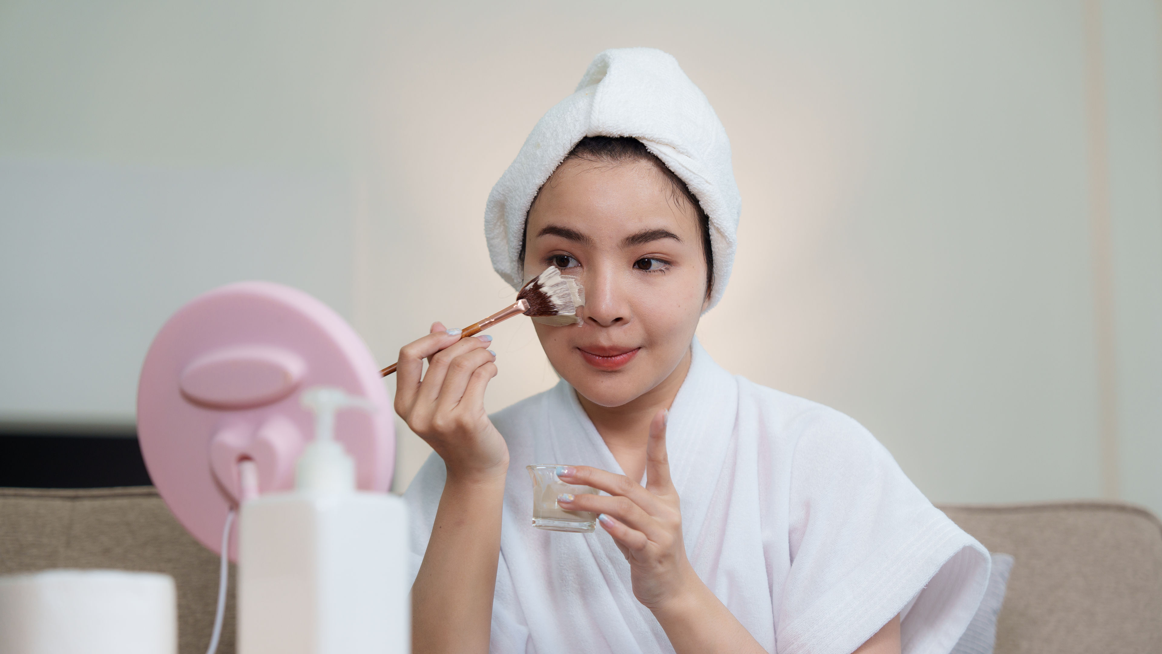 Woman in a bathrobe applying a face mask with a brush in front of a mirror.