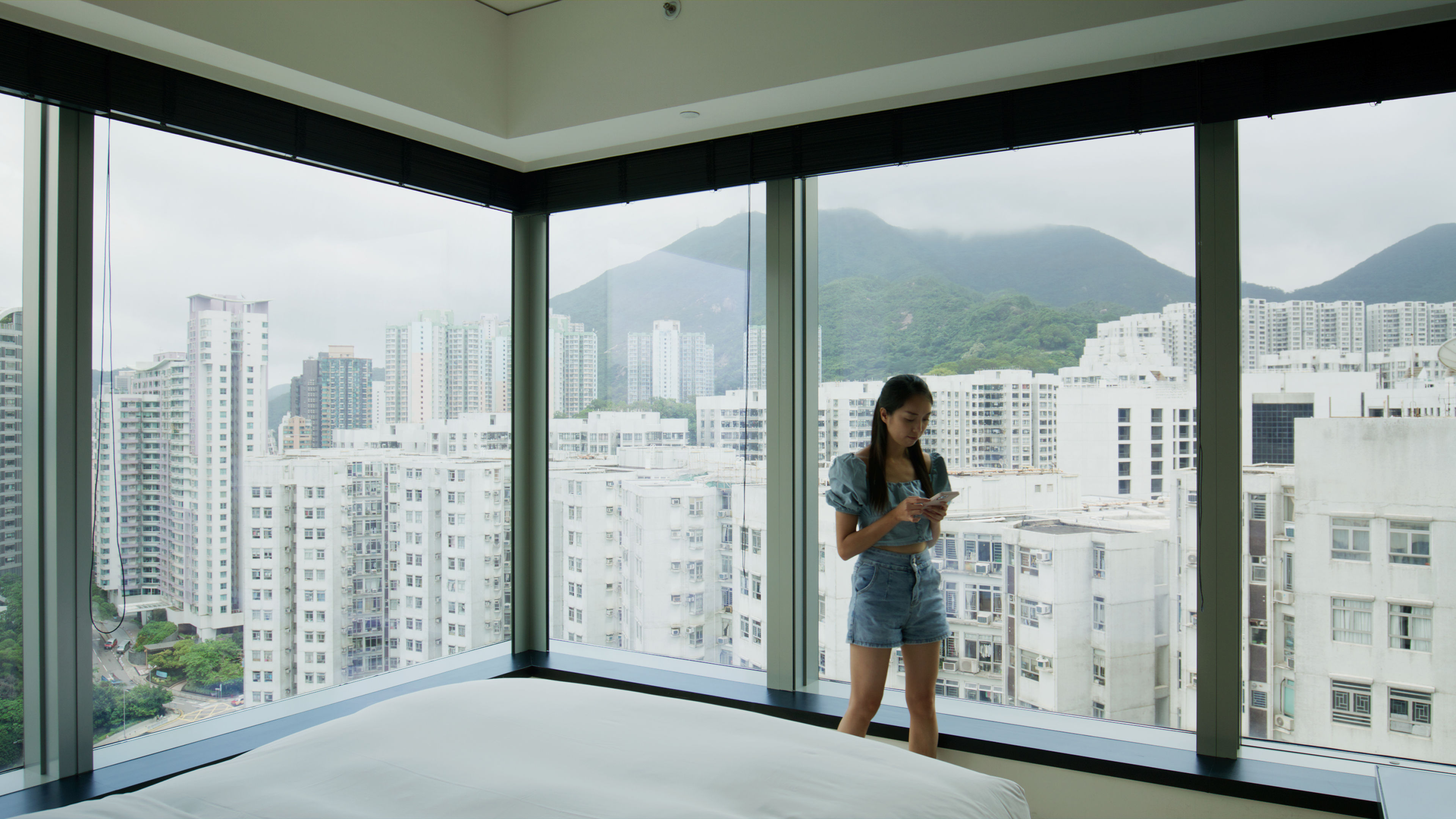 Standing in a bright room surrounded by large windows, a woman scrolls on her phone 