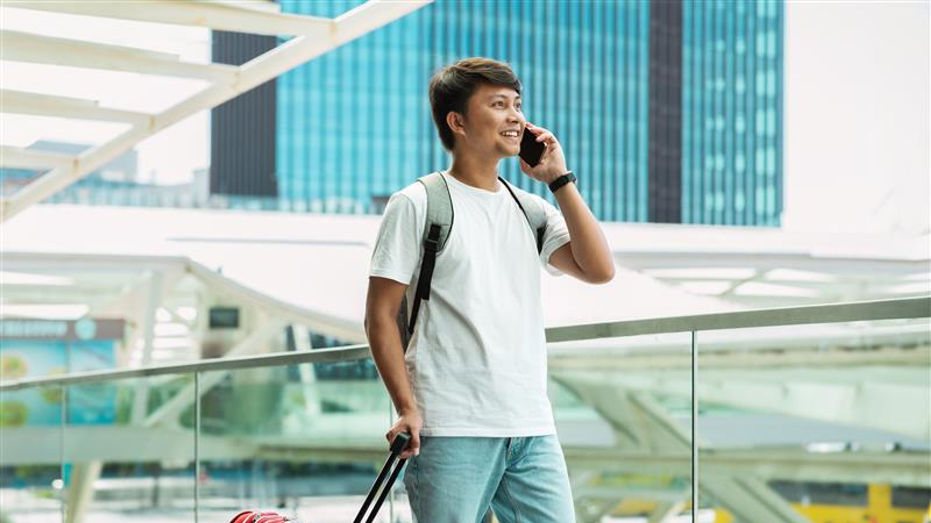 Asian guy tourist walking by airport, talking on phone, carrying red suitcase