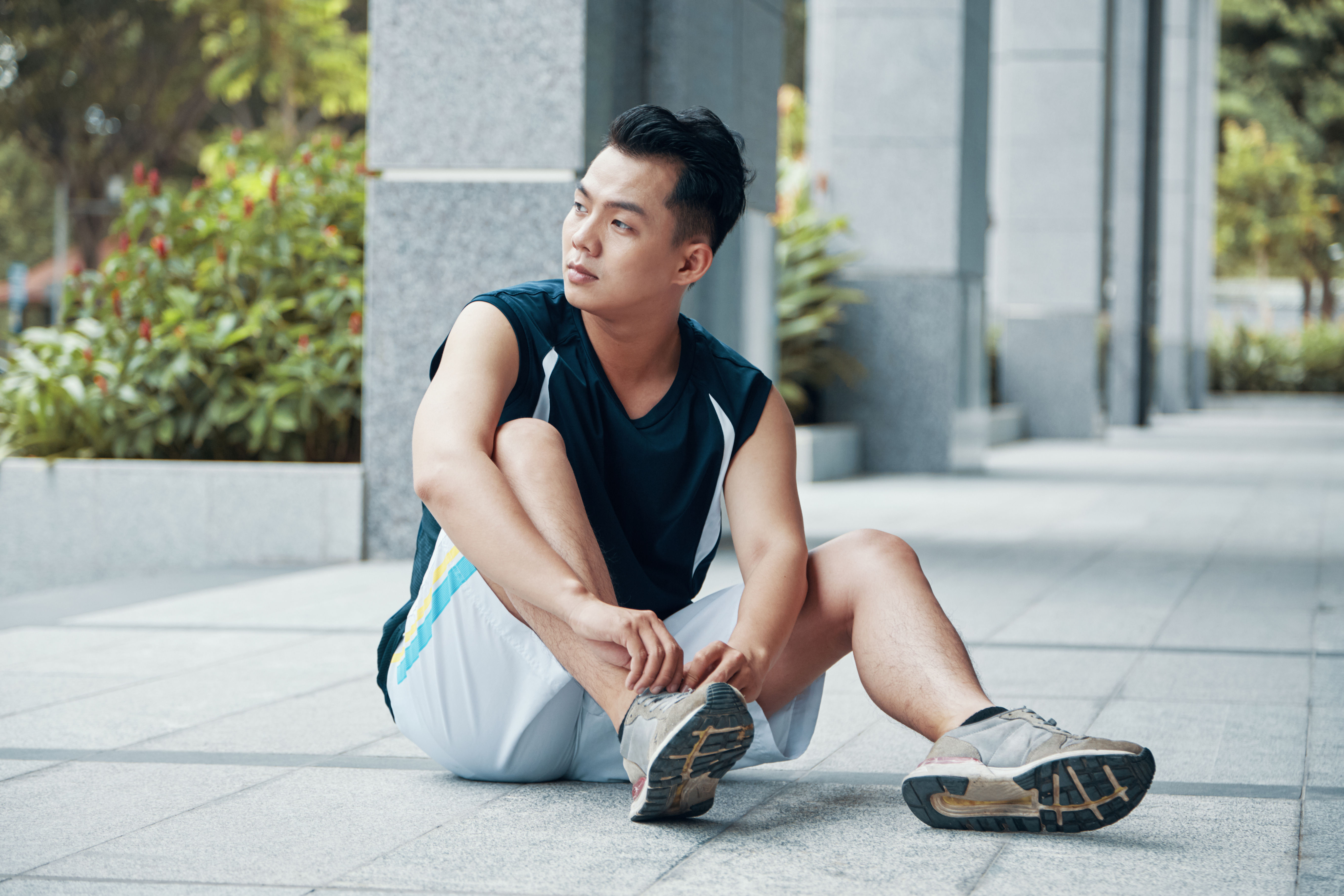 Attractive Asian guy in sportswear tying laces on sneakers and looking away while sitting on ground during outdoor training 
