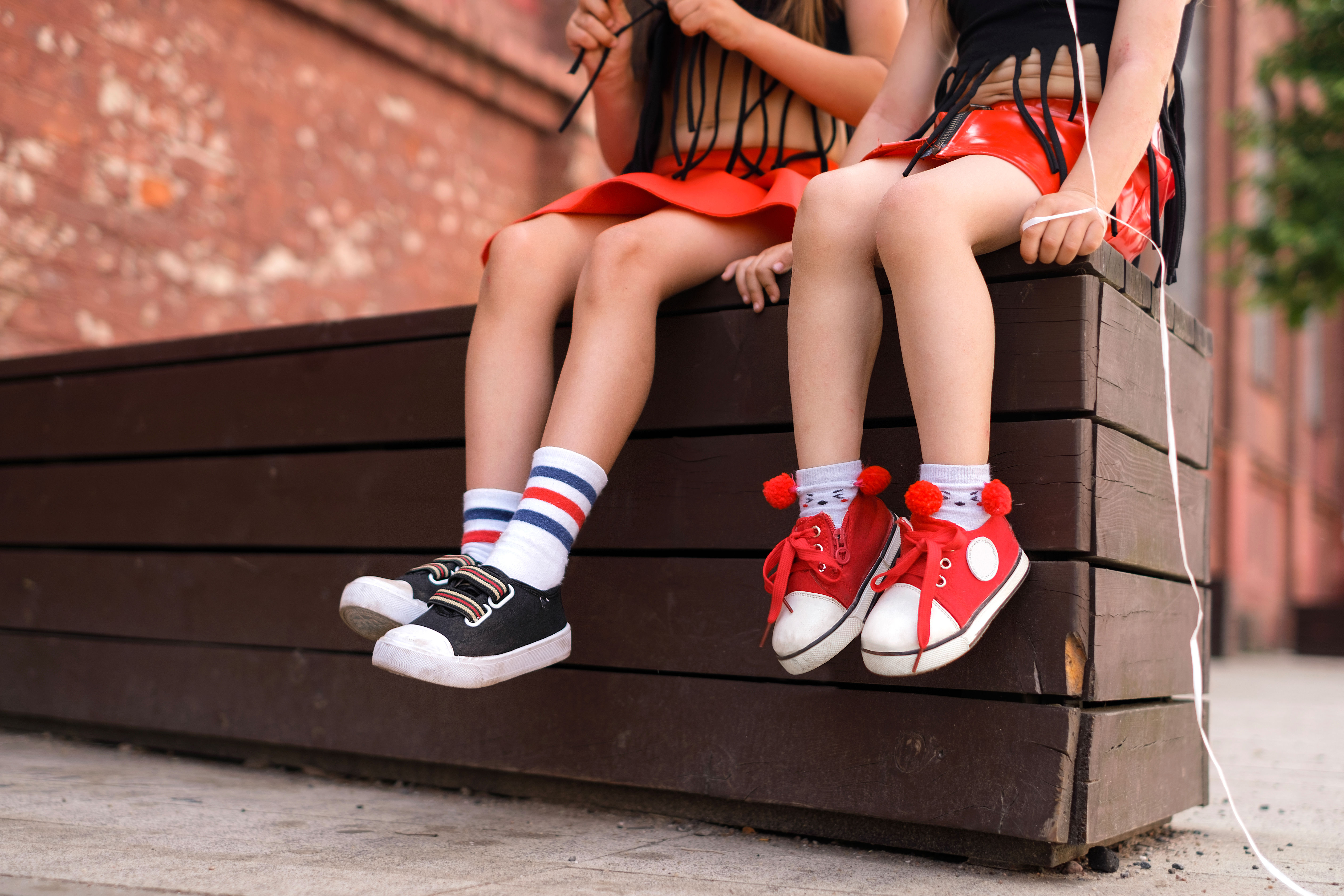 Two young girls are sitting on a wooden bench wearing colorful sneakers 
