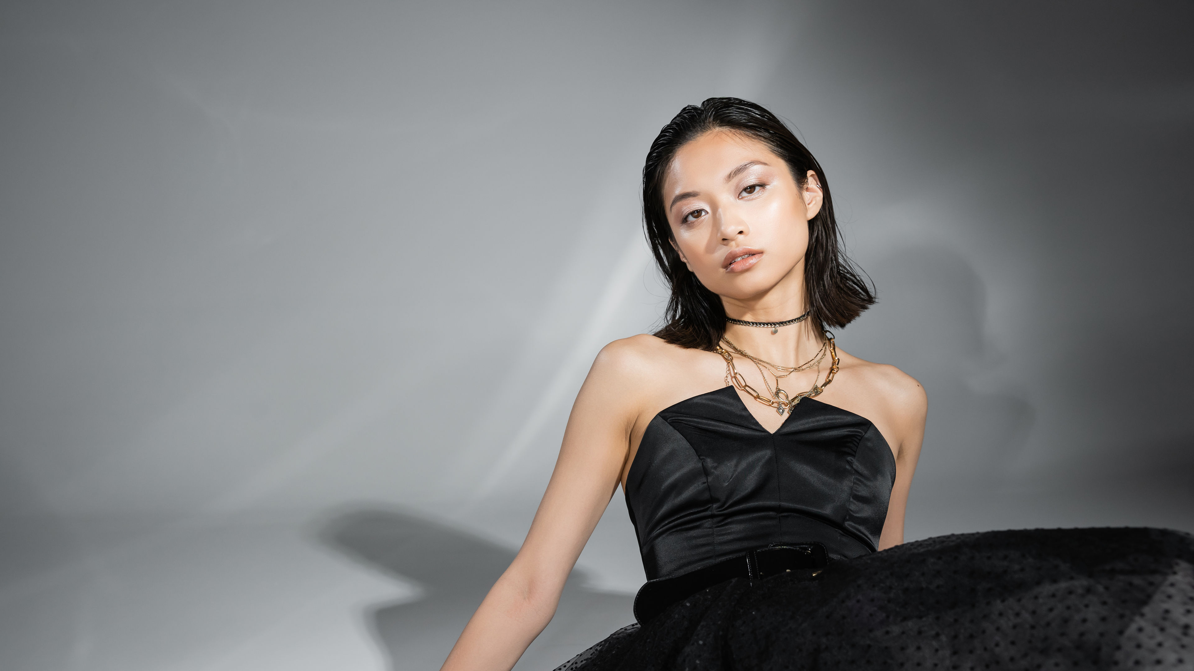 graceful asian woman with short hair sitting in black strapless dress with tulle skirt, belt while looking at camera on grey background, 