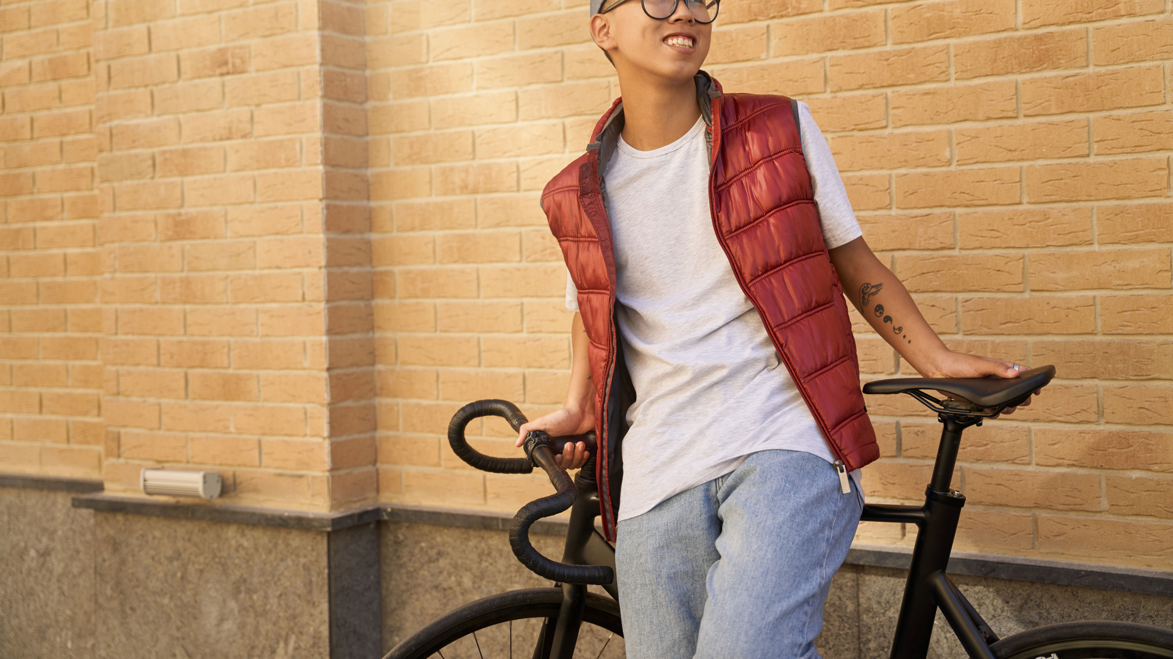 Young cheerful Asian male courier standing with his bicycle against brick wall, looking aside and smiling.