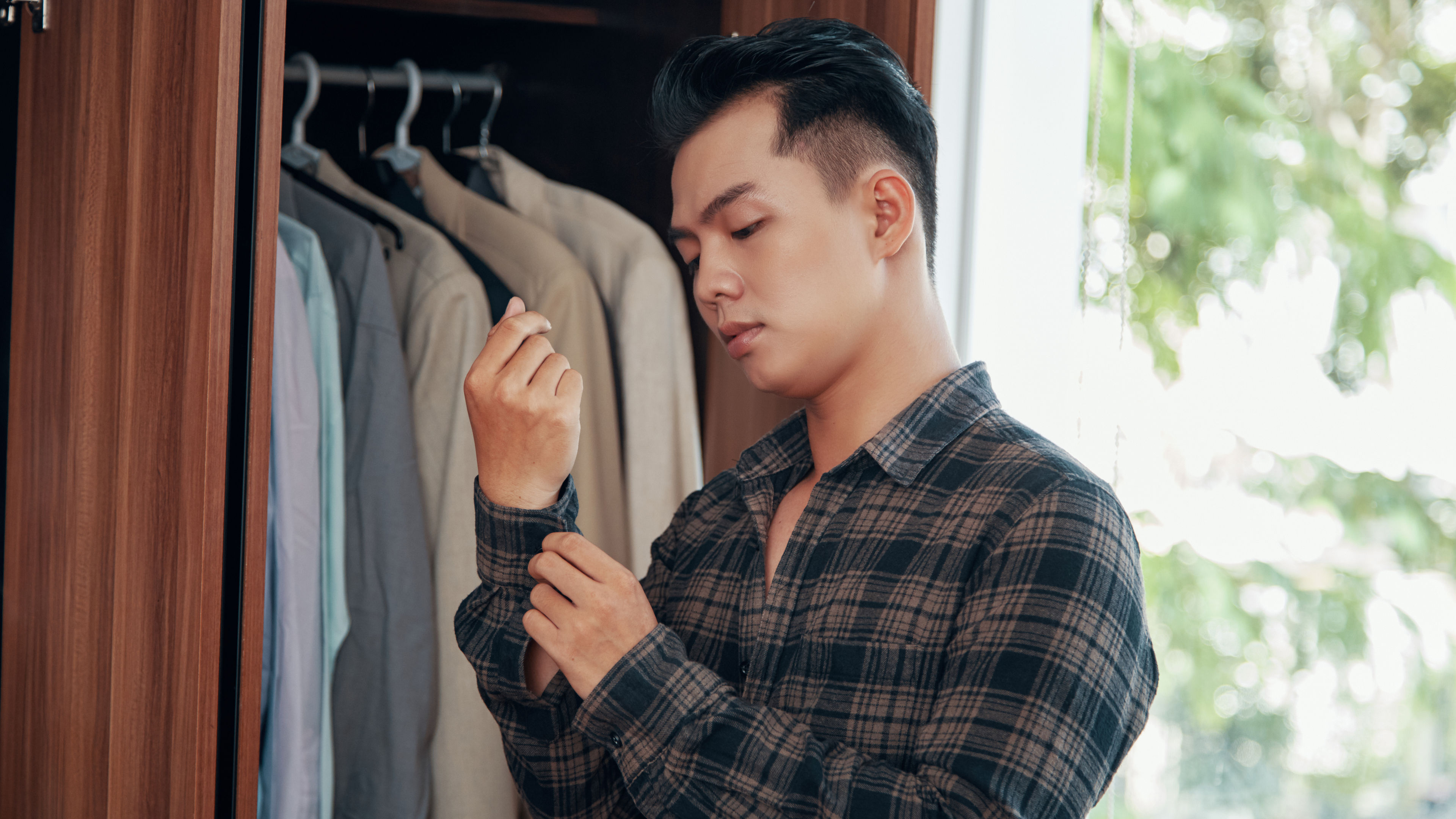 Young handsome Asian man buttoning shirt sleeves while dressing near wardrobe at home